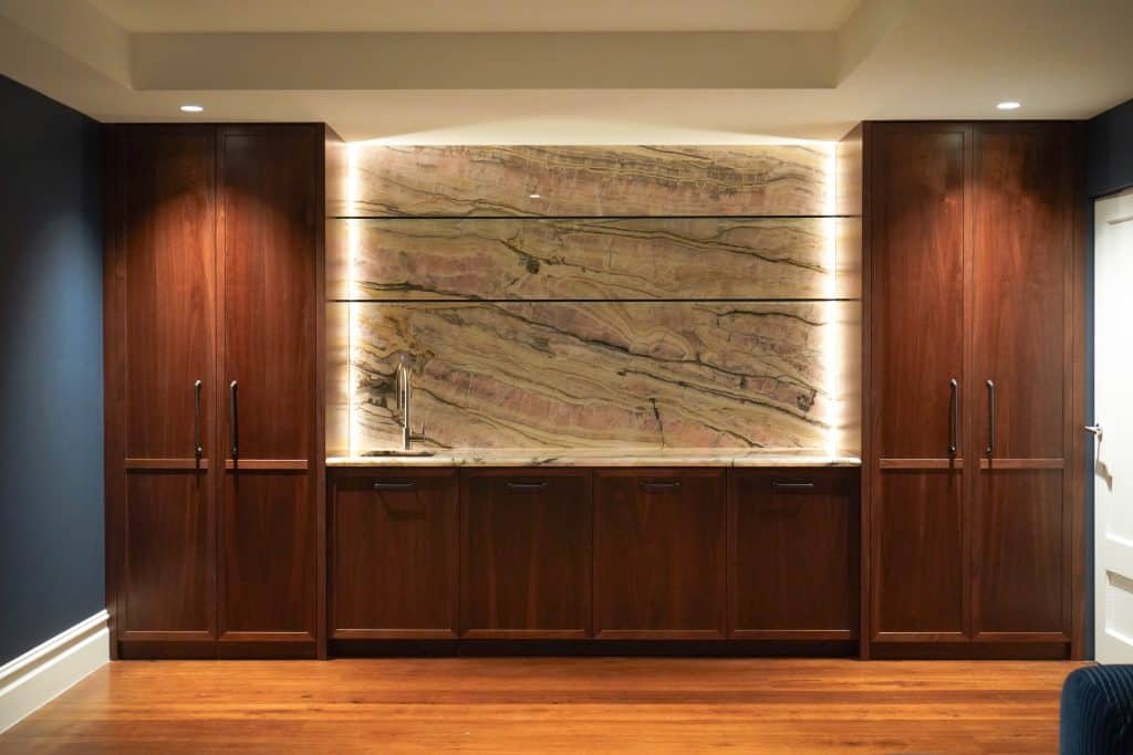 A modern home bar with dark wood cupboards, a marble splashback with prominent veining, under-cupboard lighting, and a small sink, set against a dark blue wall and wooden floor.