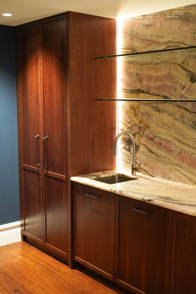 A modern kitchen corner with dark wood cupboards, a marble worktop and splashback, an undermount sink with a curved tap, glass shelves, and warm under-cupboard lighting.