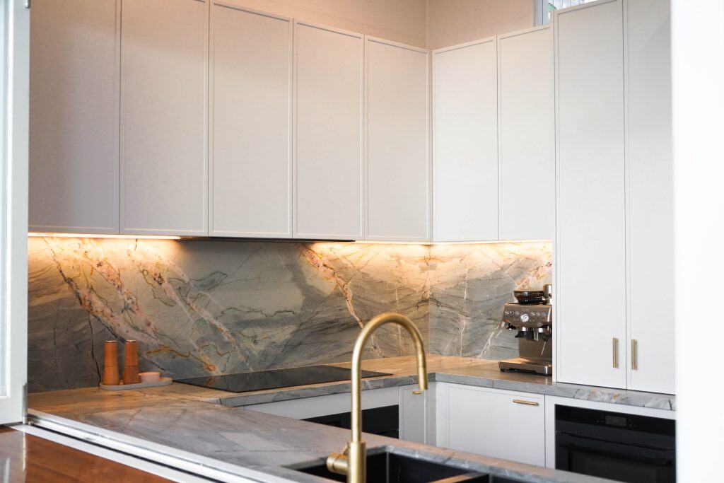 Modern kitchen with white cupboards, marble splashback and worktops, a built-in hob, a gold tap, under-cupboard lighting, and a coffee machine on the counter.