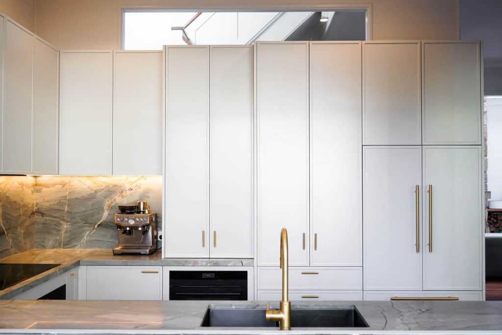 Modern kitchen with tall white cupboards, marble splashback, built-in appliances, a coffee machine on the worktop, and a gold tap over a grey marble worktop and sink.