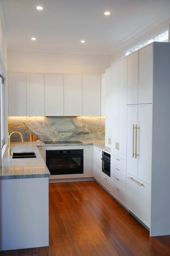 Modern kitchen with white cupboards, gold handles, marble splashback, built-in oven, and wooden flooring. Under-cupboard lighting highlights the worktops and splashback. A gold tap sits on the left worktop.