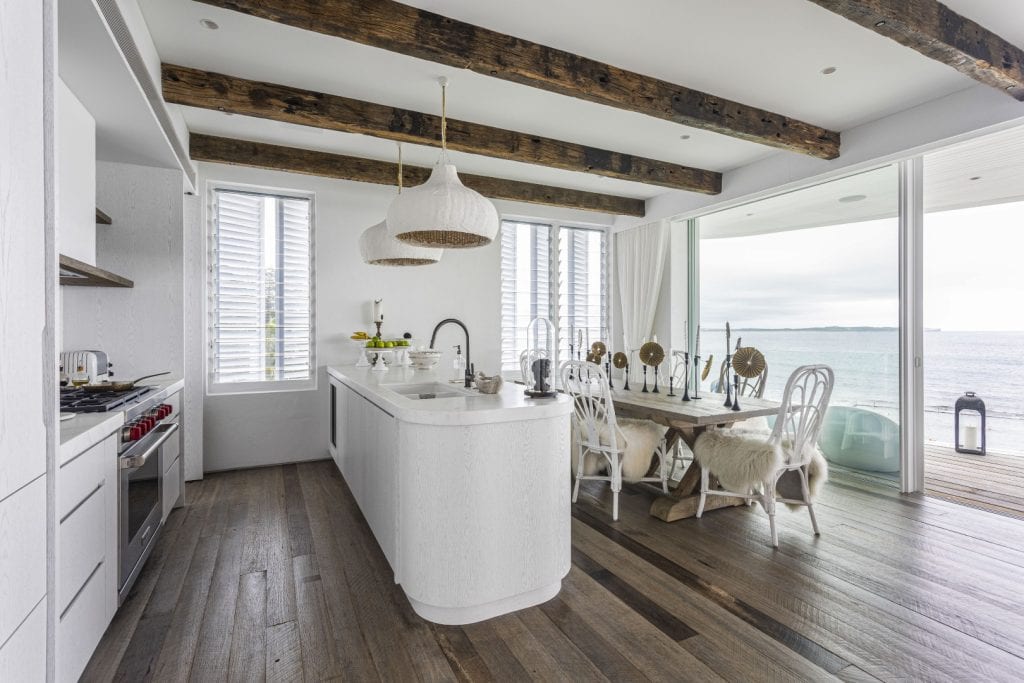 Bright, modern kitchen with white cupboards, a large island, rustic wooden beams, and wooden floors. Large windows and sliding doors offer a sea view. Dining table with white chairs and natural accents is set for a meal.