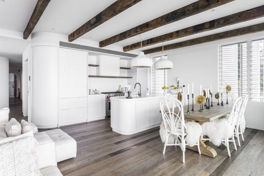 A modern, bright kitchen and dining area with white cupboards, wooden ceiling beams, pendant lights, and a rustic dining table with white chairs and fluffy seat covers on a wooden floor.