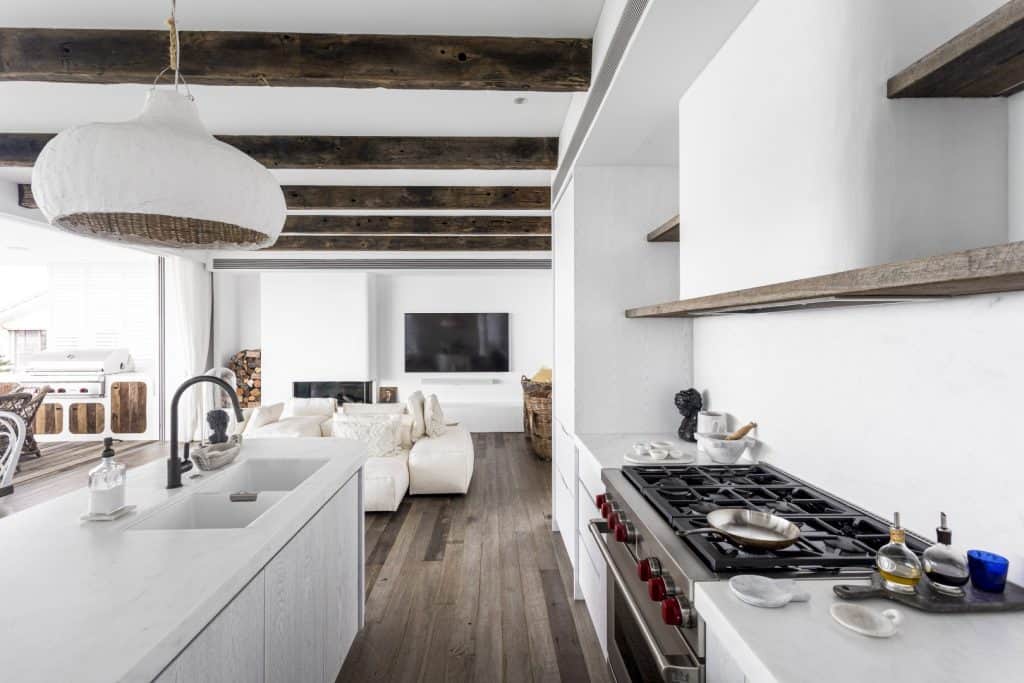 Modern kitchen with white worktops and rustic wooden beams, featuring a gas cooker, open shelving, and sink island. In the background, a cosy living area with a cream sofa and flat-screen TV is visible.