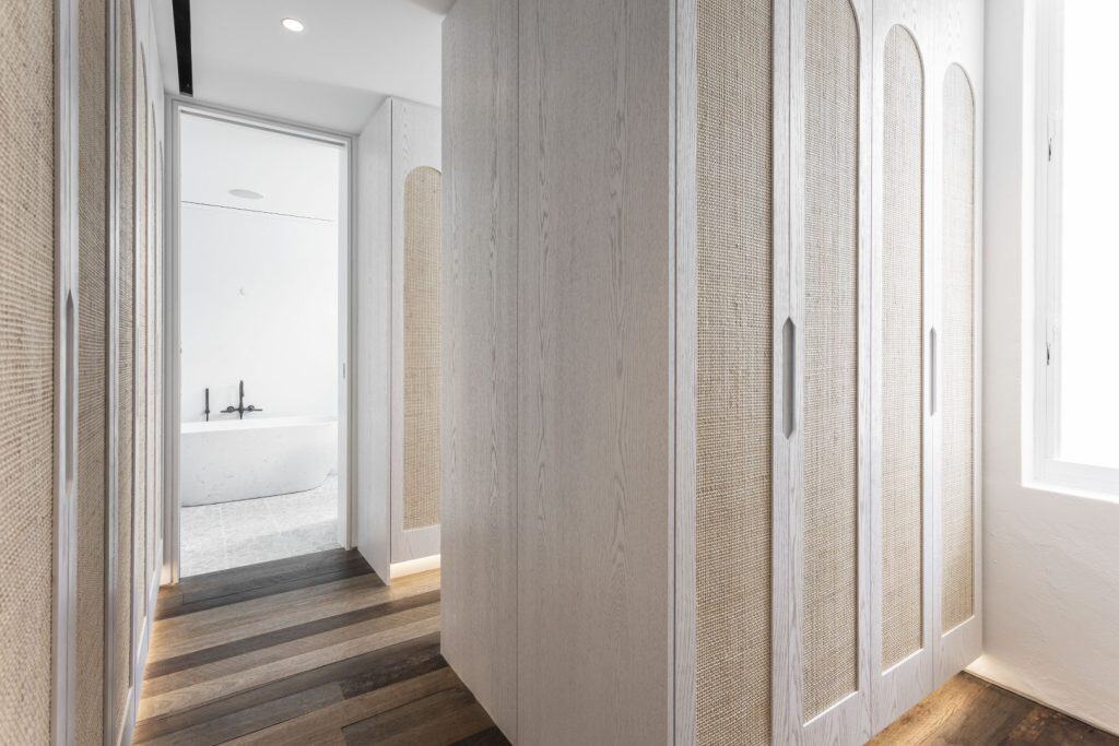 A modern hallway with light wood wardrobes featuring arched details and textured panels, leading to a bright bathroom with a white freestanding bath and black fittings. Sunlight enters through a window on the right.