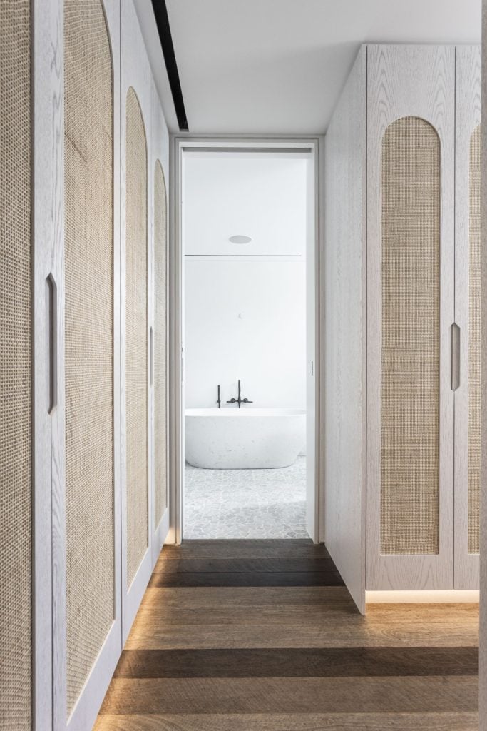 A corridor with light wood cupboards featuring woven panels leads to a minimalist white bathroom with a freestanding bath and black fittings. The floor is dark wood with a striped pattern.