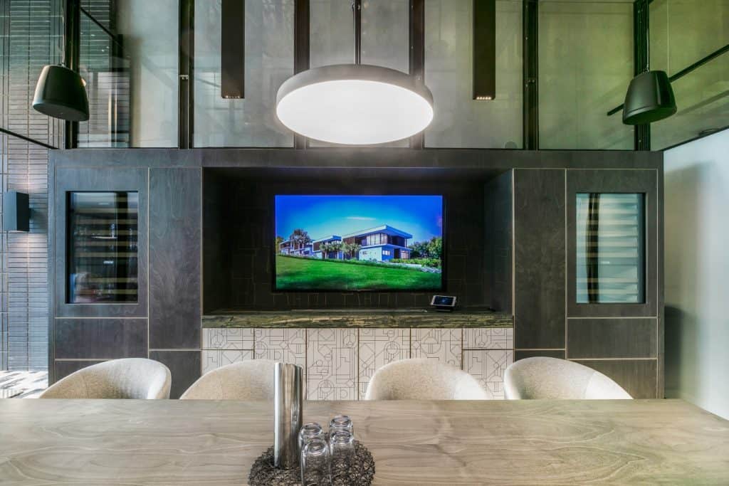 Modern conference room with a large TV displaying a house, surrounded by dark cupboards and shelves. A round light hangs above a wooden table set with glasses, a metal vase, and decorative items.