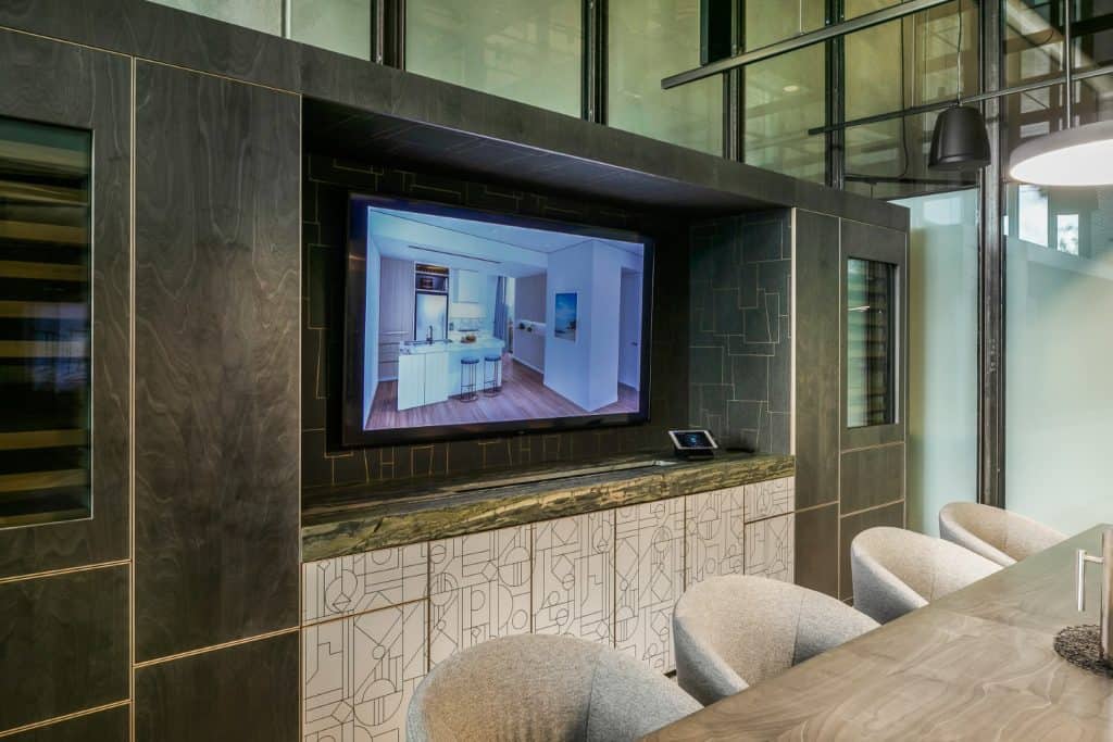 A modern conference room features a large wall-mounted screen displaying a kitchen image, surrounded by dark wood panelling, with a marble-topped cabinet and grey upholstered chairs at a table.