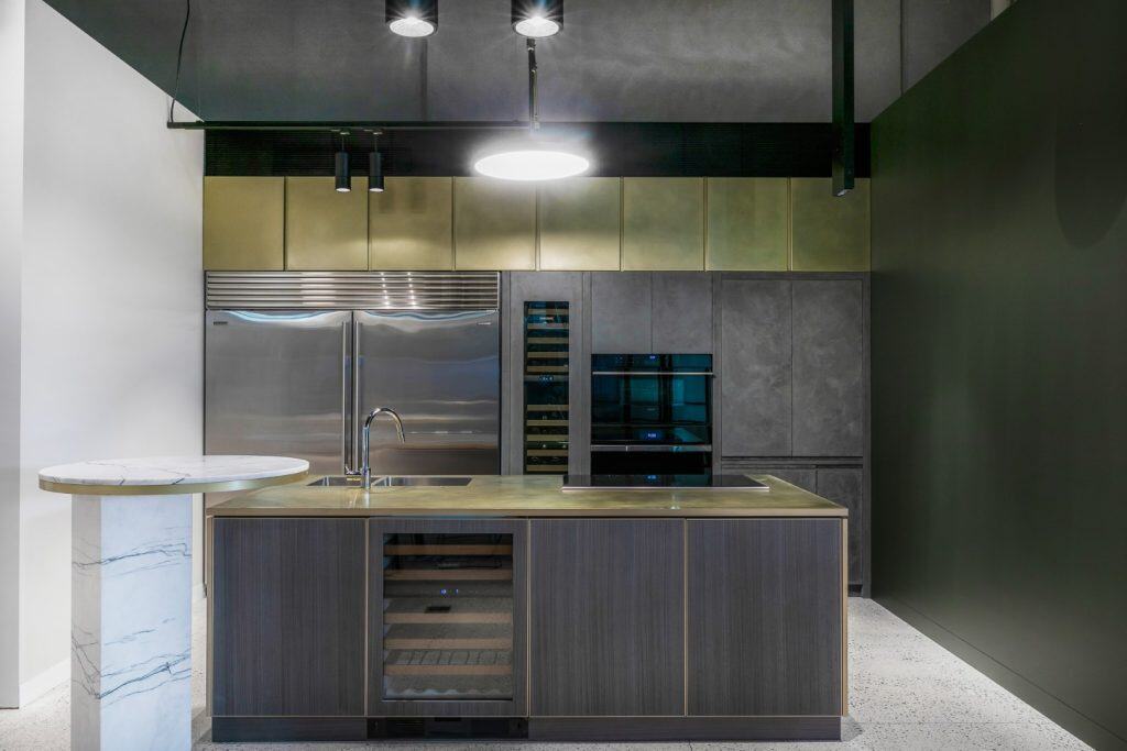 Modern kitchen with a central island featuring a built-in wine cooler and sink, metallic cupboards, double oven, large stainless steel fridge, and a round marble-topped table extension.