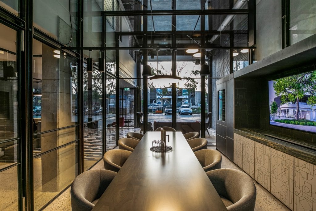 A modern meeting room with a long, dark table surrounded by grey chairs, glass walls, overhead lighting, and a large TV screen on the right displaying an outdoor scene. Outside, parked cars and trees are visible through the glass.