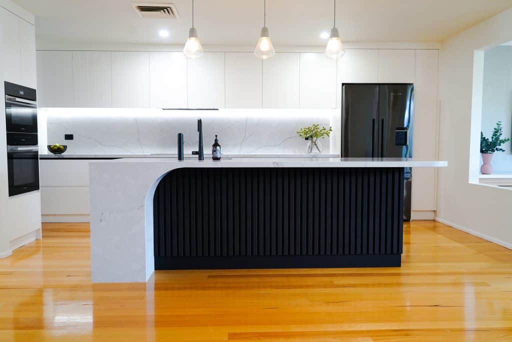 Modern kitchen with a large black and white island, marble worktop, black fridge, built-in ovens, pendant lights, and wooden flooring. Minimalist decor with a small vase of greenery and a bowl of fruit.