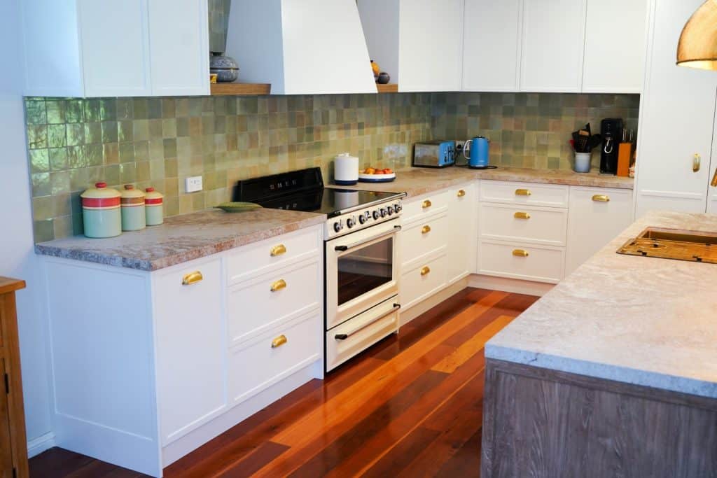 A modern kitchen with white cupboards, gold handles, light stone worktops, a stainless steel cooker, various small appliances, green-tiled splashback, and polished wooden floors.