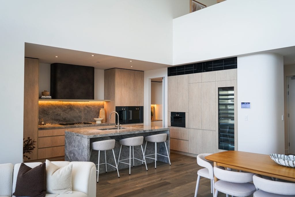 Modern kitchen with wooden cupboards, a marble island with three bar stools, built-in appliances, open shelves, and under-cupboard lighting; adjacent to a dining table with white chairs and a sofa in the foreground.
