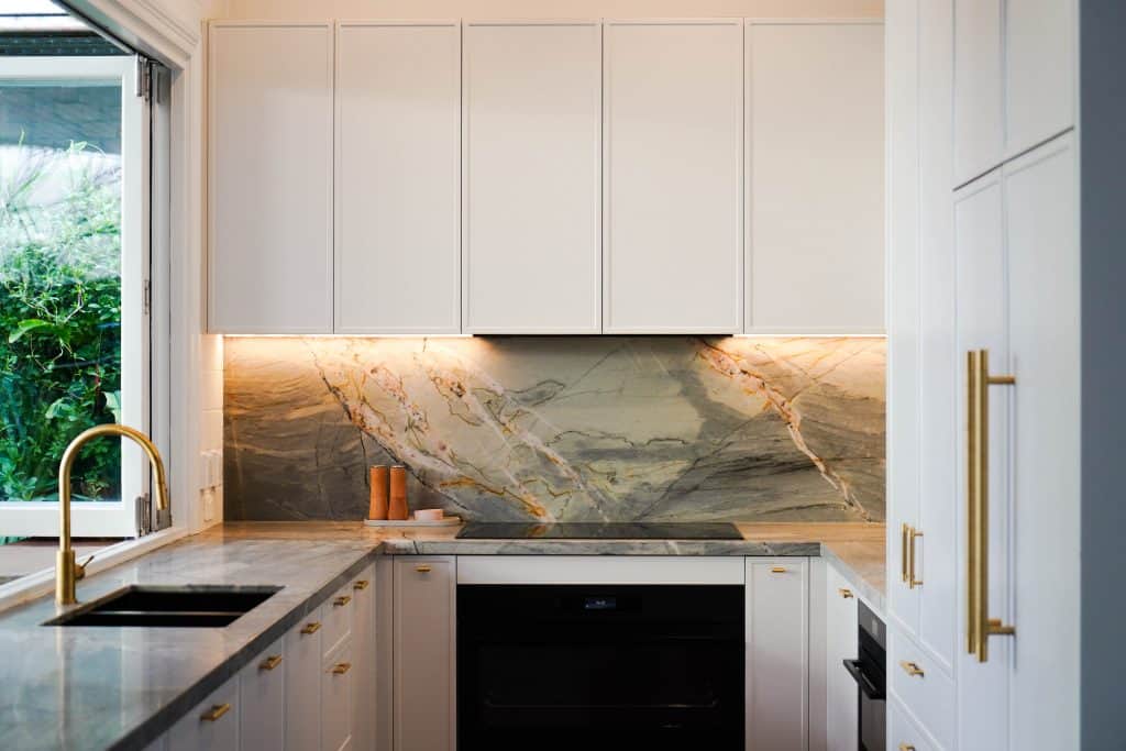 Modern kitchen with white cupboards, gold fittings, marble splashback, grey worktops, black oven, built-in hob, and a gold tap next to a large window showing greenery outside. Warm under-cupboard lighting is on.