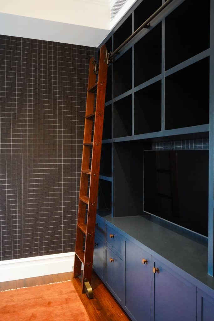 A dark wooden ladder leans against built-in blue shelving with cupboards and open cubbies. The shelves stand beside a wall covered in black grid-patterned wallpaper. A rust-coloured rug is on the floor.