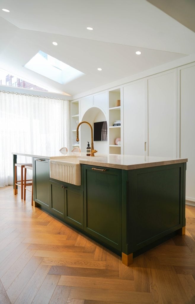 A modern kitchen with white cupboards, a green island featuring a farmhouse sink and gold tap, wooden herringbone floors, and large windows with sheer curtains letting in natural light.