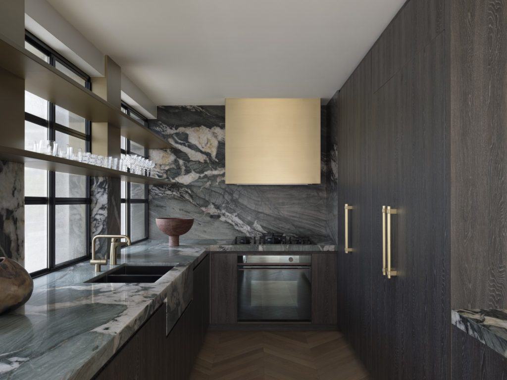 Modern kitchen with dark wood cabinets, marble worktops and splashback, gold fittings, open shelves with glassware, built-in oven, and large gold cooker hood under wide windows.