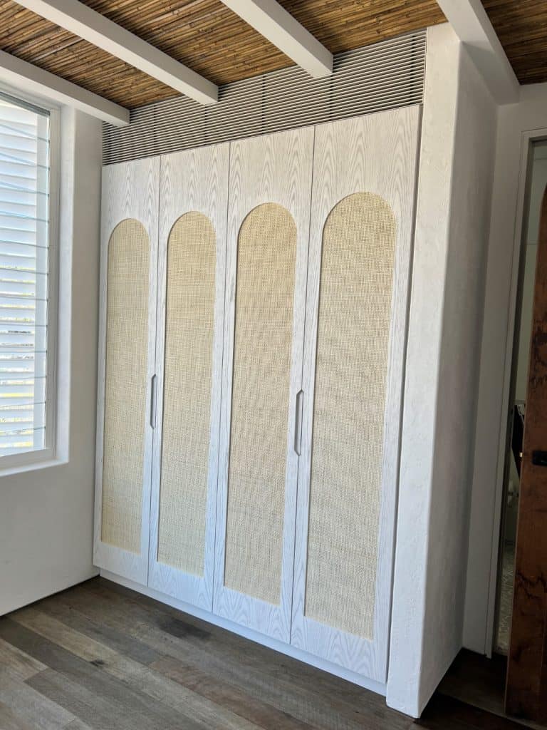 A white fitted wardrobe with four arched panel doors and subtle handles stands against a white wall, next to a window with blinds. The floor has wooden planks, and the ceiling features exposed beams and bamboo.