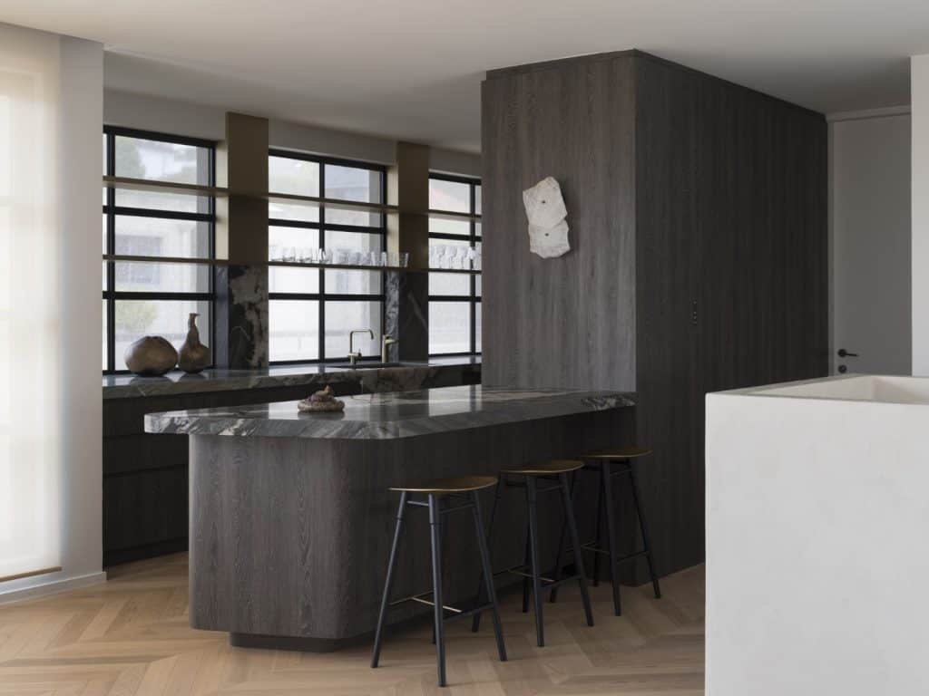 A modern kitchen with dark wood cabinets, a marble worktop island, three black bar stools, large windows with black frames, and minimalist decor including vases and a wall-mounted sculpture.