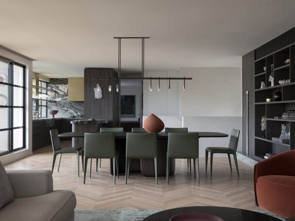 Modern dining area with a dark oval table, green chairs, a terracotta centrepiece, open shelving, large windows, chevron wood flooring, and a marble-accented kitchen in the background.